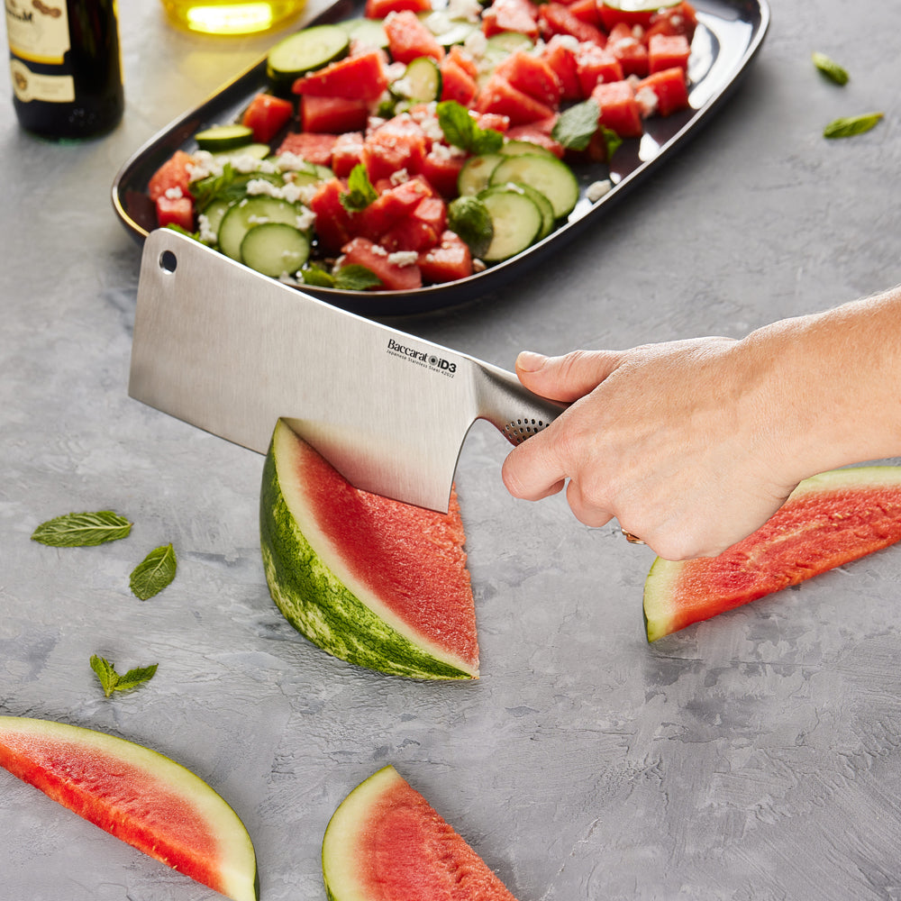 Person slicing watermelon with a cleaver next to a salad on a gray countertop - Baccarat
