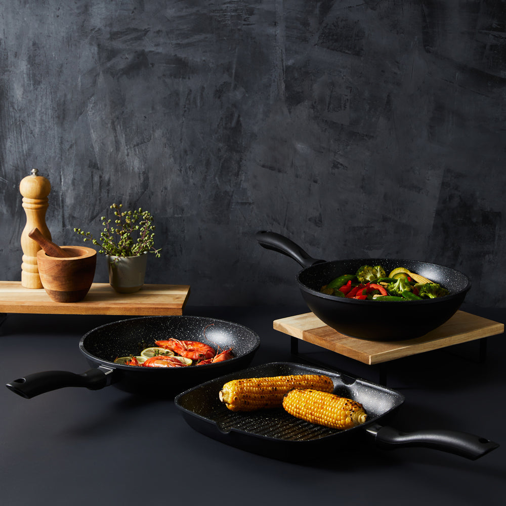 Three frying pans and a grill pan on a dark counter with wooden utensils and herbs - Baccarat