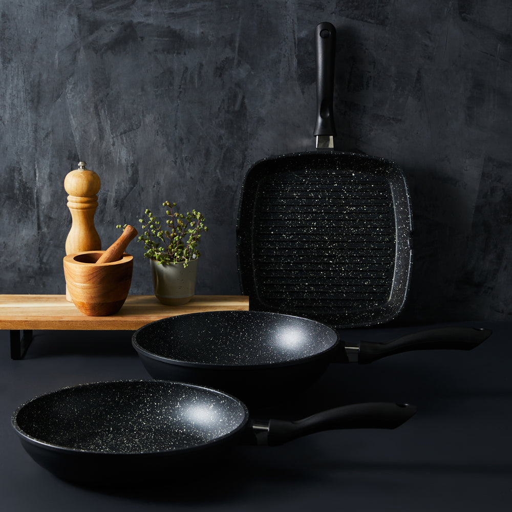 Three frying pans and a grill pan on a dark counter with wooden utensils and herbs - Baccarat