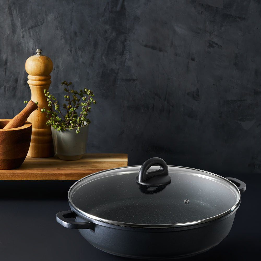 The covered black sauté pan resting on a countertop with a pepper mill, mortar and pestle, and potted plant in the background against a dark wall.