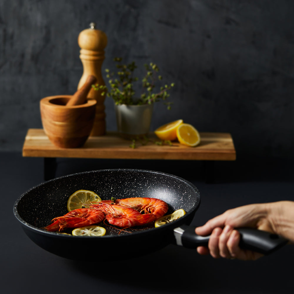 A hand holds a frying pan with grilled salmon and lemon slices stone grey - Baccarat