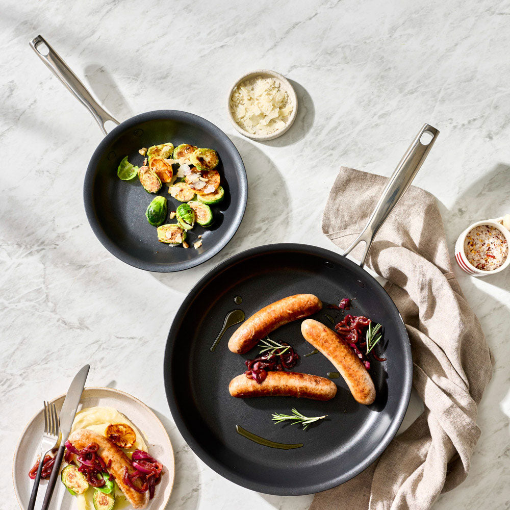 An overhead shot shows two frying pans with food on a table setting alongside a cloth and small bowls.