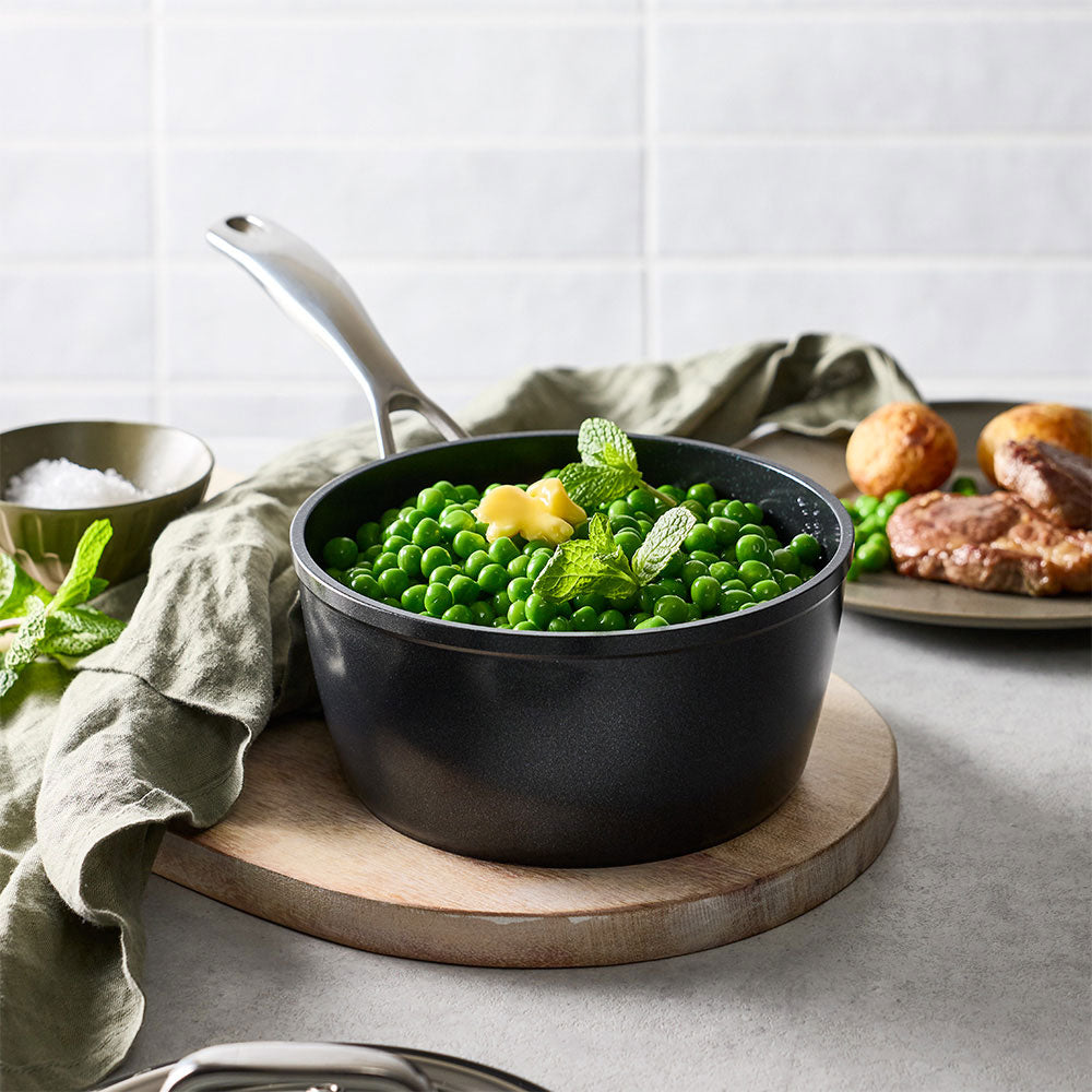 The black saucepan is filled with green vegetables and placed on a wooden board on a kitchen counter.