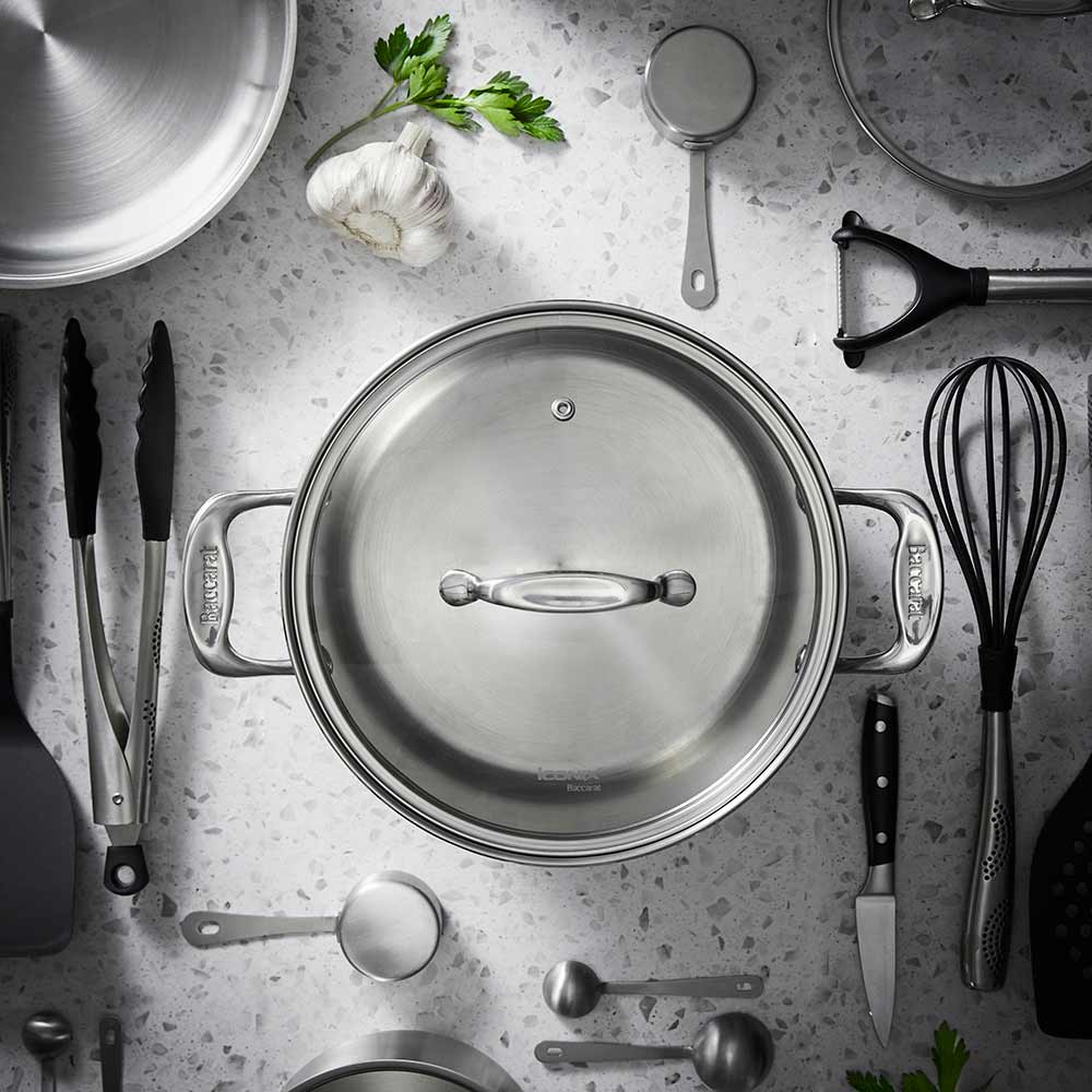 A silver stockpot with lid surrounded by kitchen utensils on a gray background - Baccarat