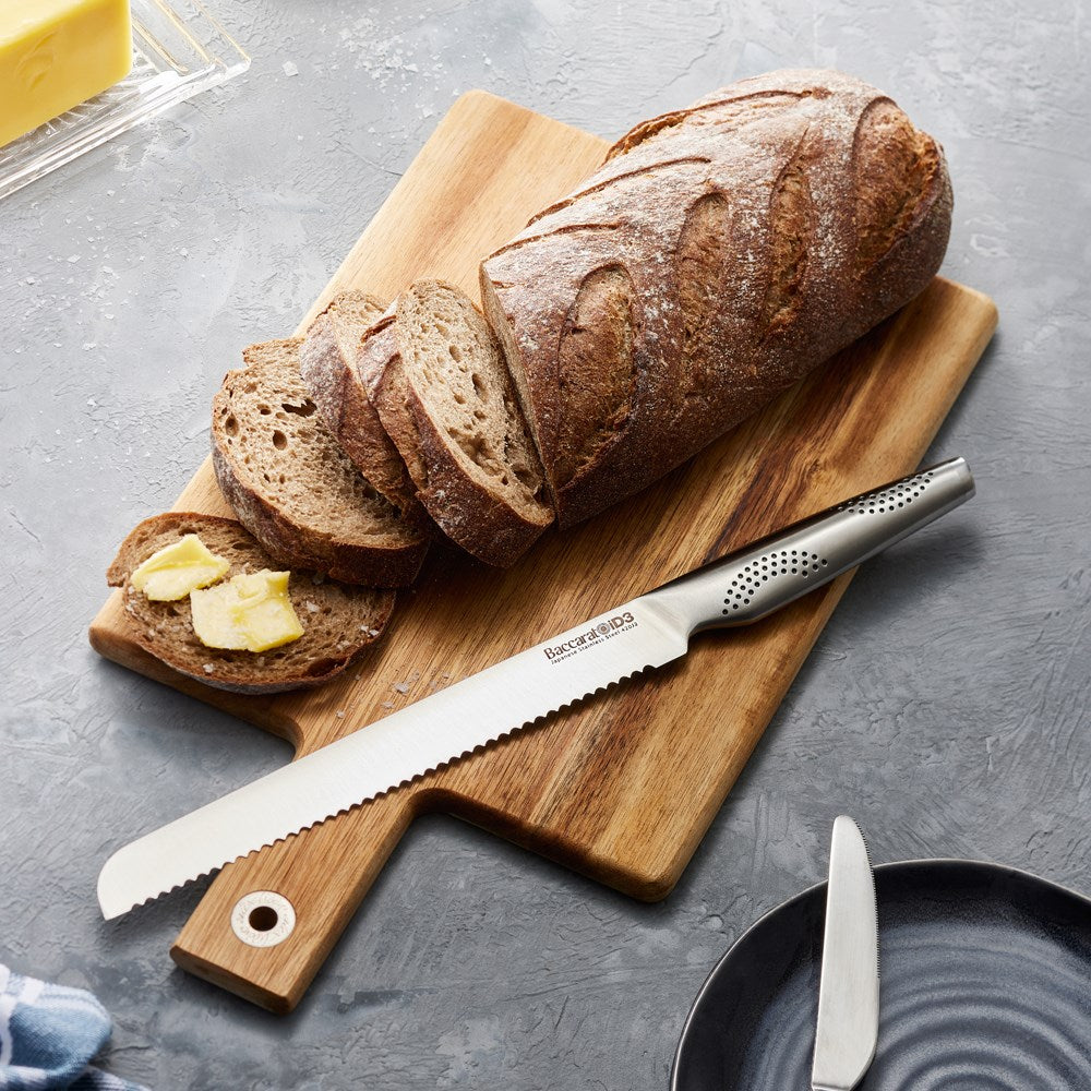A sleek serrated bread knife is positioned on a wooden cutting board next to fresh bread - Baccarat