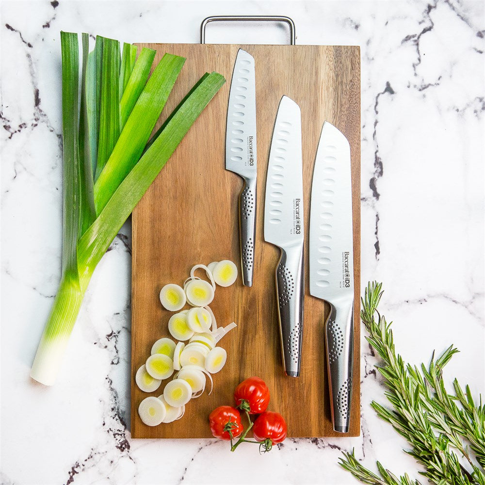 Three santoku knives resting on a wooden cutting board with fresh herbs and vegetables - Baccarat