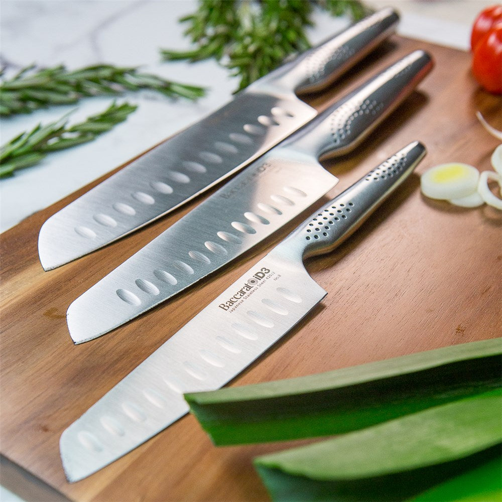 Three santoku knives resting on a wooden cutting board with fresh herbs and vegetables - Baccarat