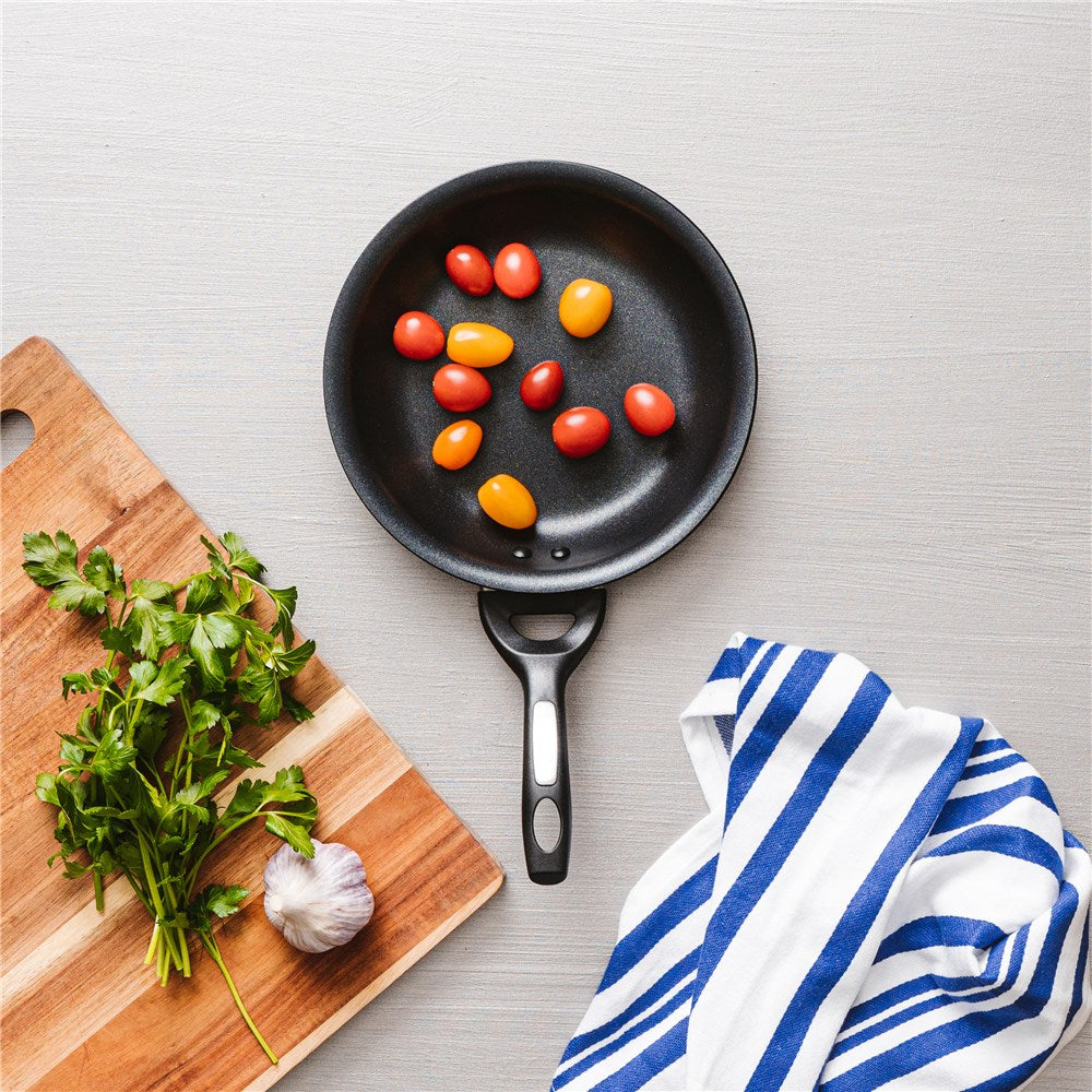 A black frying pan with vegetables including green peppers and tomatoes on a grey surface - Baccarat