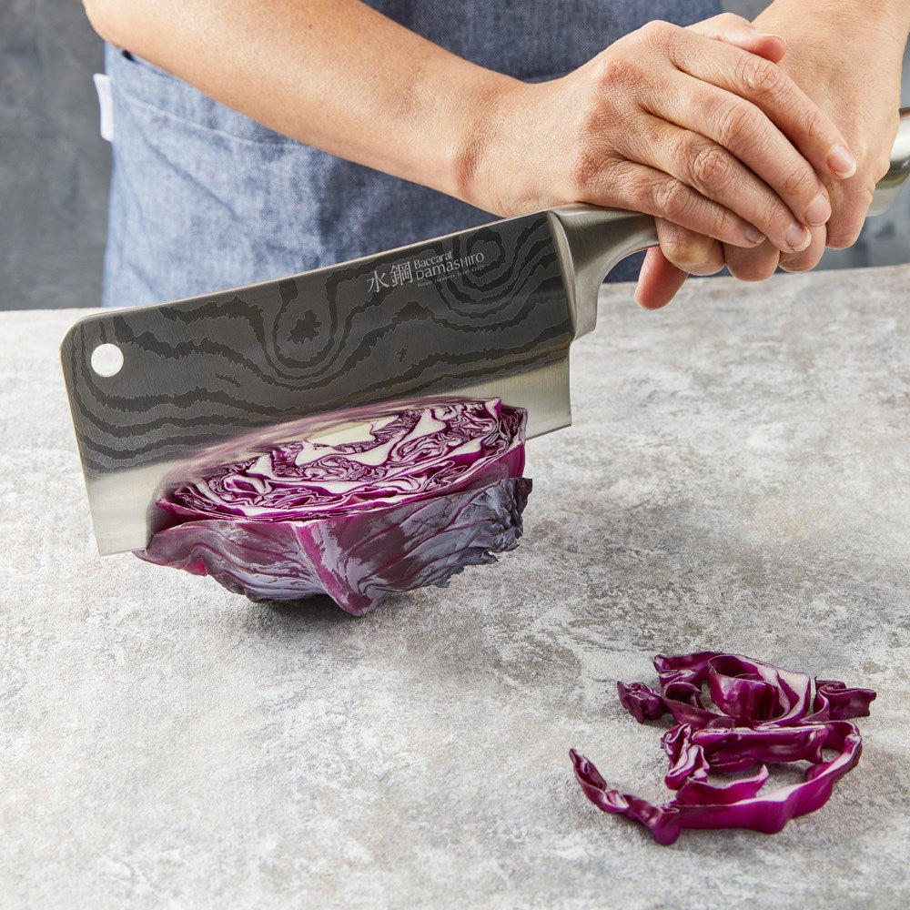 A Baccarat Damashiro cleaver with a sleek finish on a rustic surface surrounded by fresh ingredients