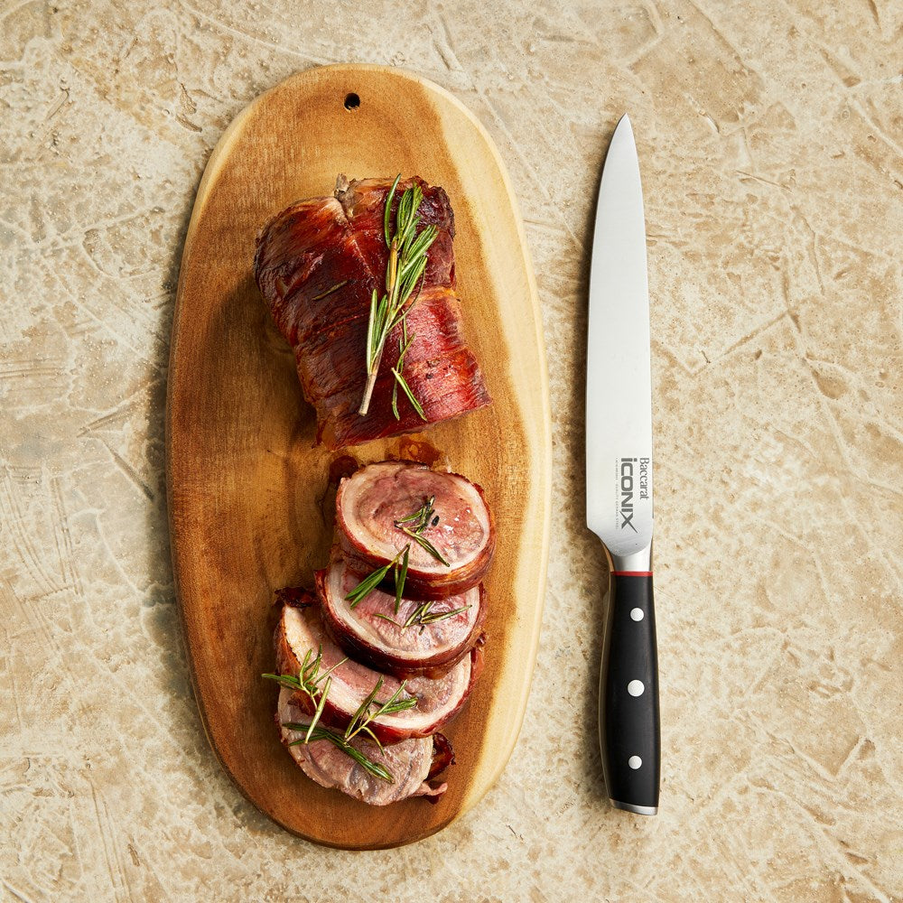 Sliced meat on a wooden board garnished with herbs next to a Baccarat Iconix Carving Knife 20cm