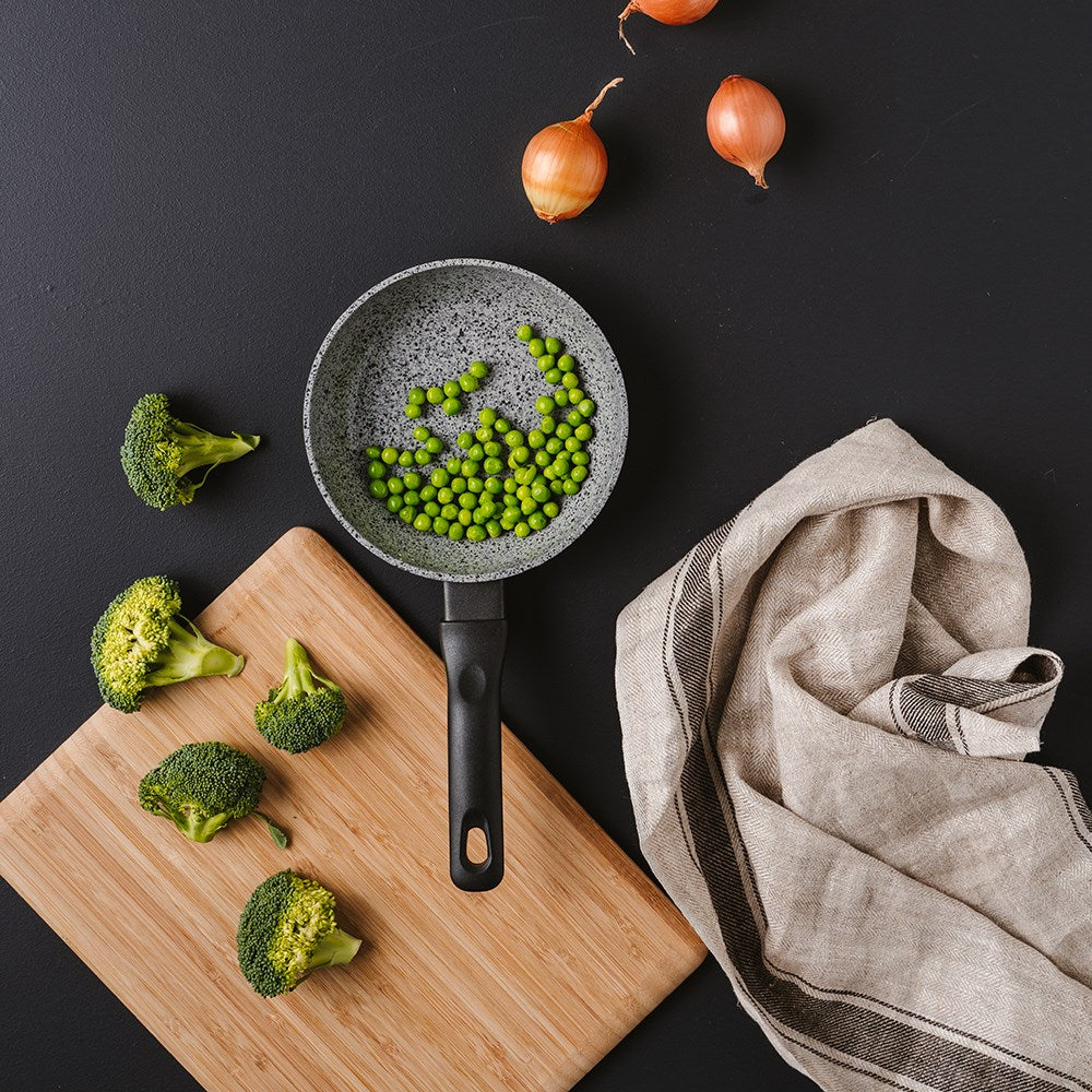 Frying pan with green peas broccoli and onions on a black surface beside a beige cloth - Baccarat
