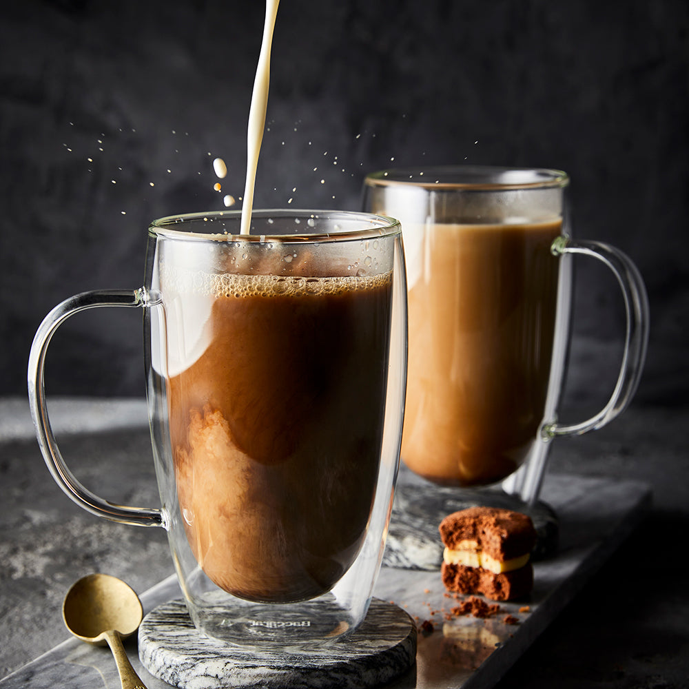 Two glass mugs of creamy iced coffee being poured against a dark background red casserole - Baccarat