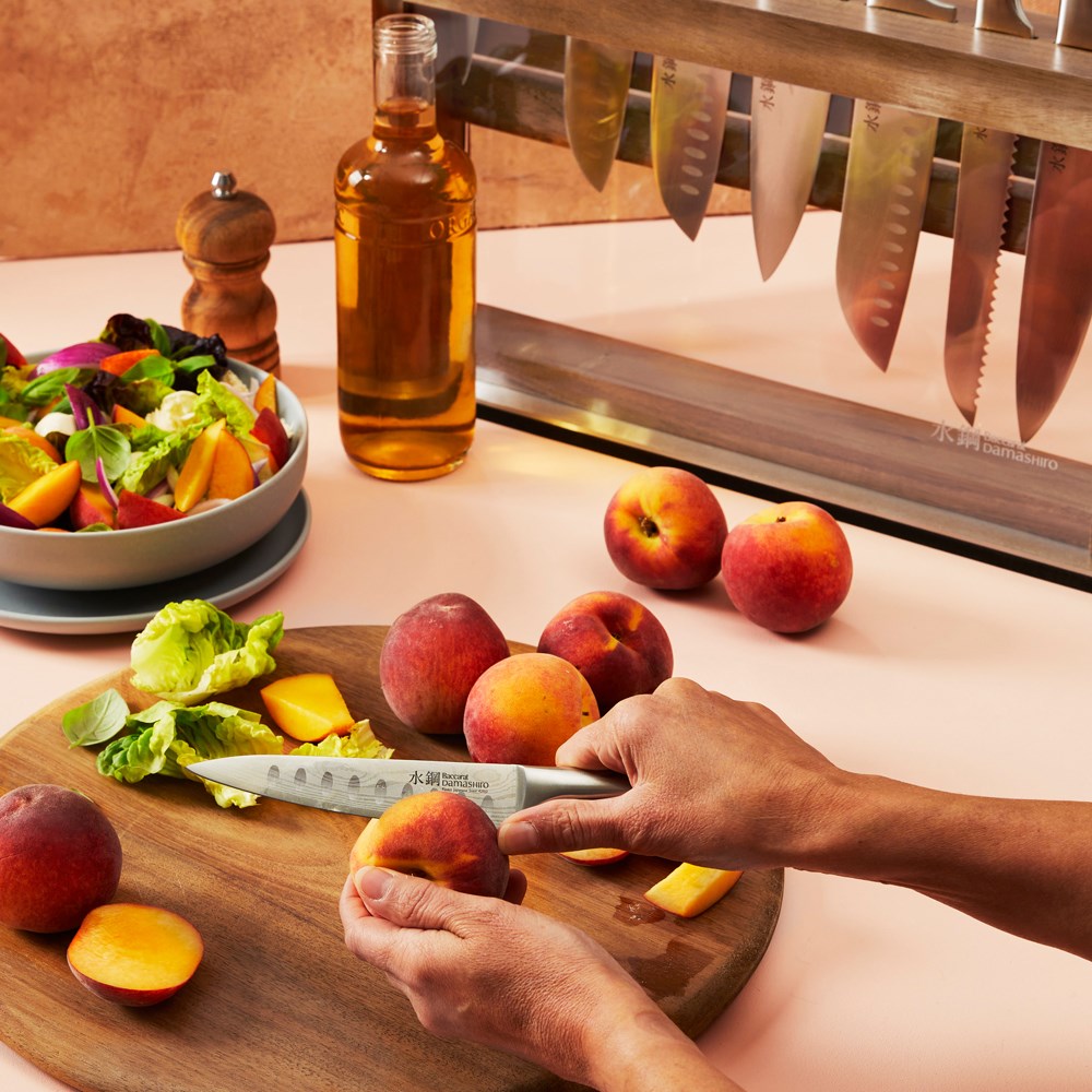 Slices of peaches on a wooden board next to a salad bowl and knives in the background - Baccarat