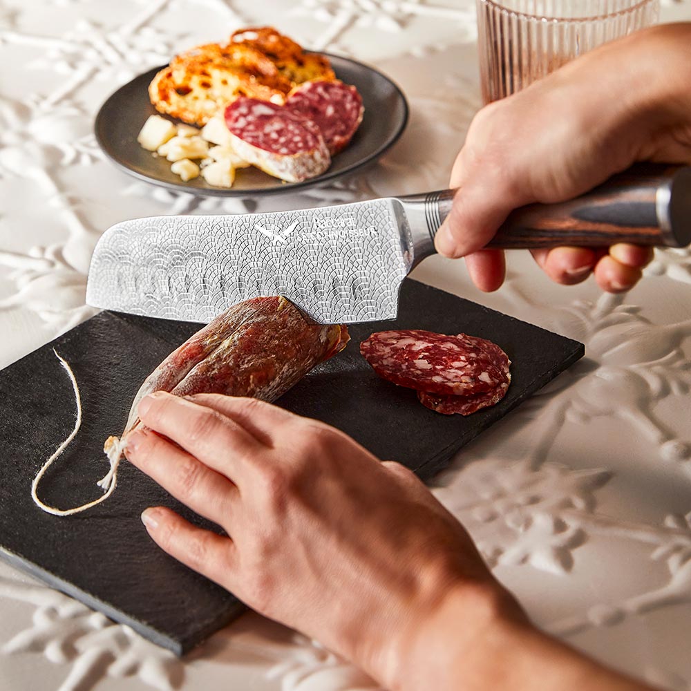 Hands slicing cured sausage on a black slate board metal knife food - Santoku knife 14cm - Baccarat