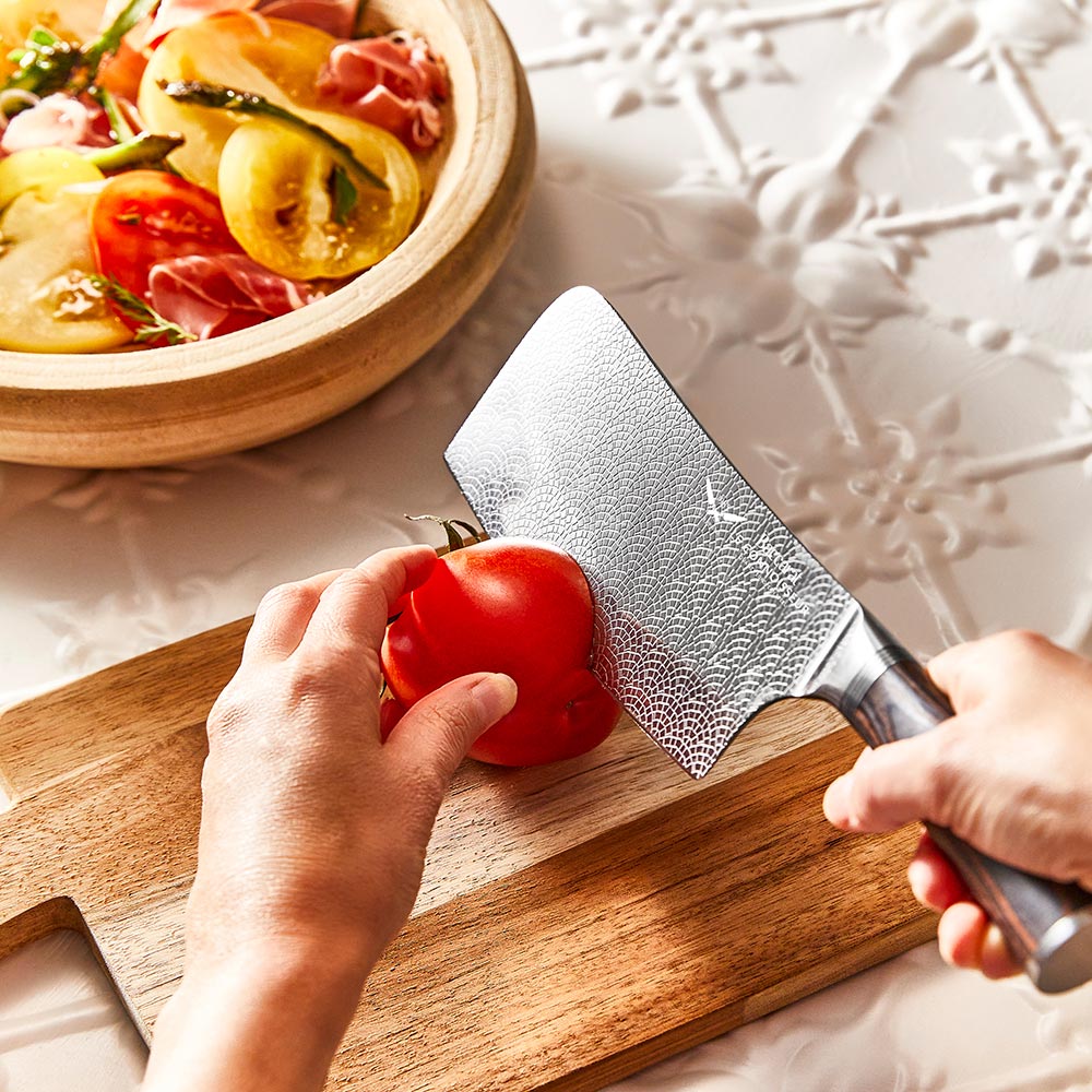 A hand slicing a red tomato with silver cleaver on a wooden cutting board - Baccarat Le Connoisseur