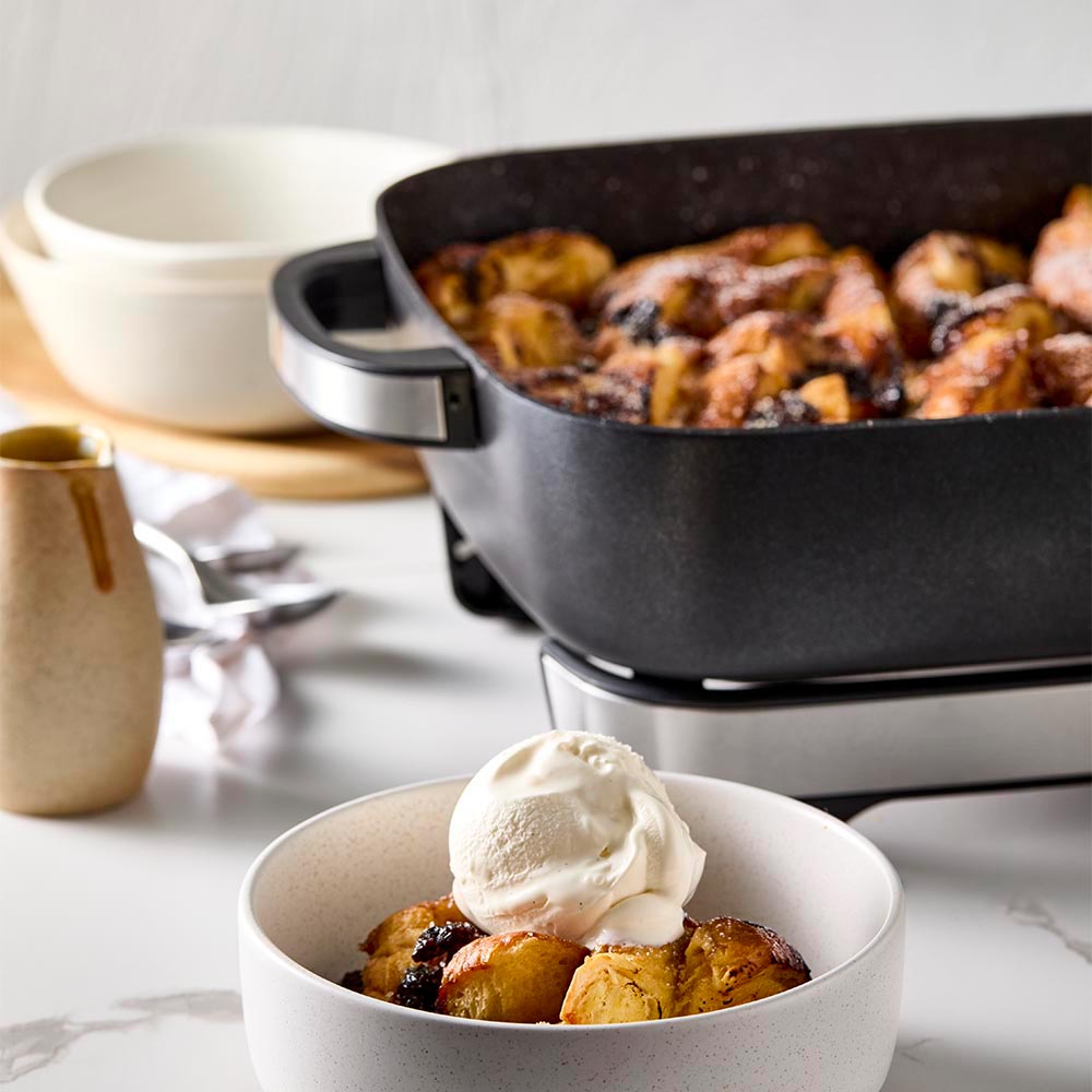 A black pan of bread pudding with a bowl of ice cream on a white countertop black frypan - Baccarat