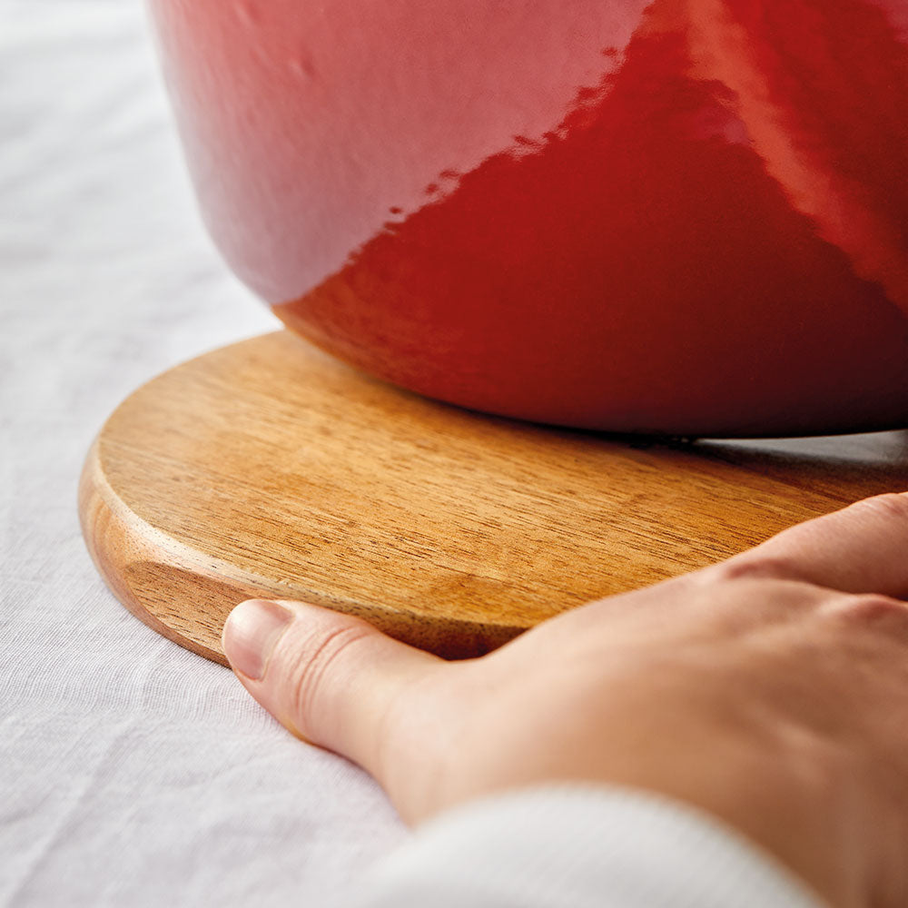 A hand gently holding a wooden trivet under a shiny red pot on a white surface red pot - Baccarat