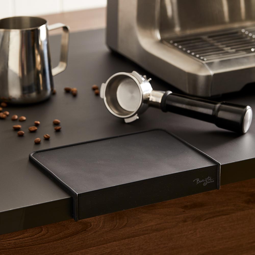 A coffee tamper and portafilter on a Baccarat Barista Brillante Coffee Mat beside scattered beans