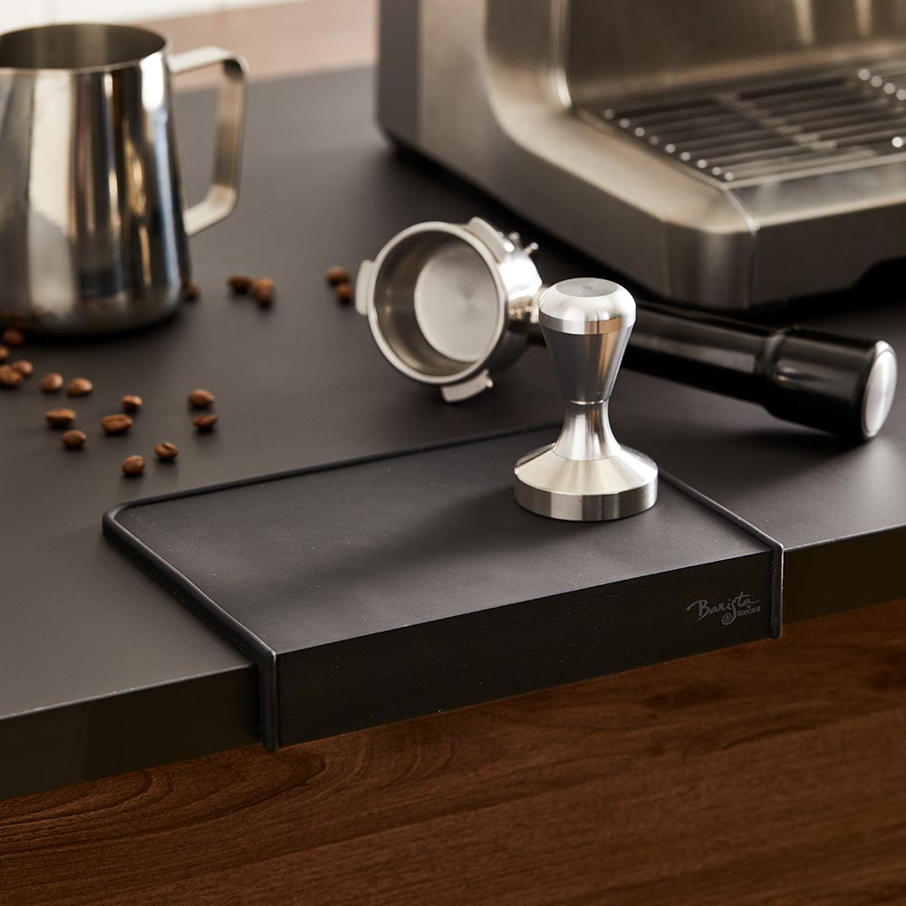 A coffee tamper and portafilter on a Baccarat Barista Brillante Coffee Mat beside scattered beans