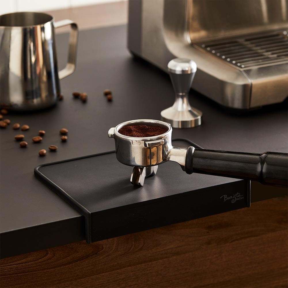 A coffee tamper and portafilter on a Baccarat Barista Brillante Coffee Mat beside scattered beans