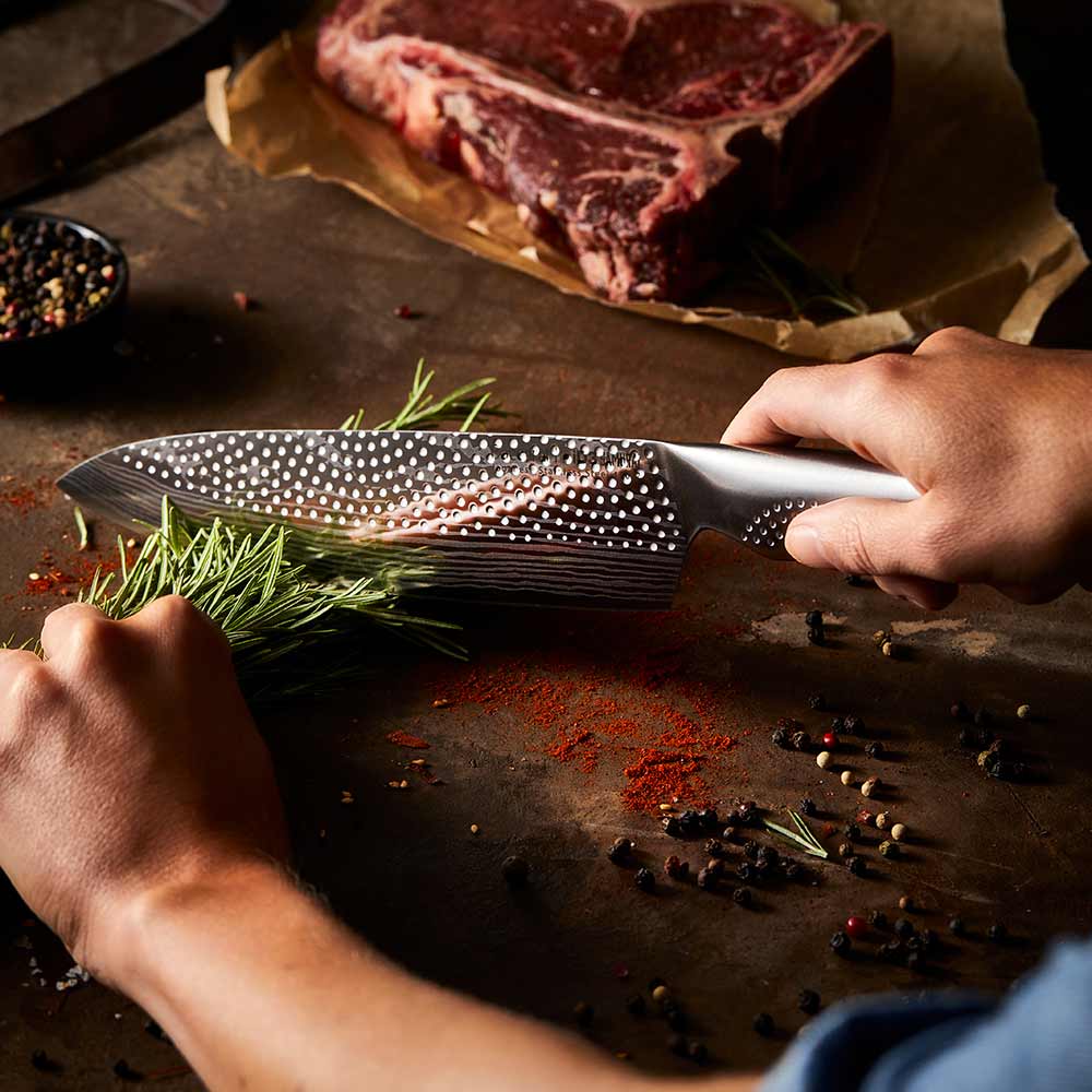 A person chops herbs in a kitchen with large steak in background on a wooden board - Baccarat