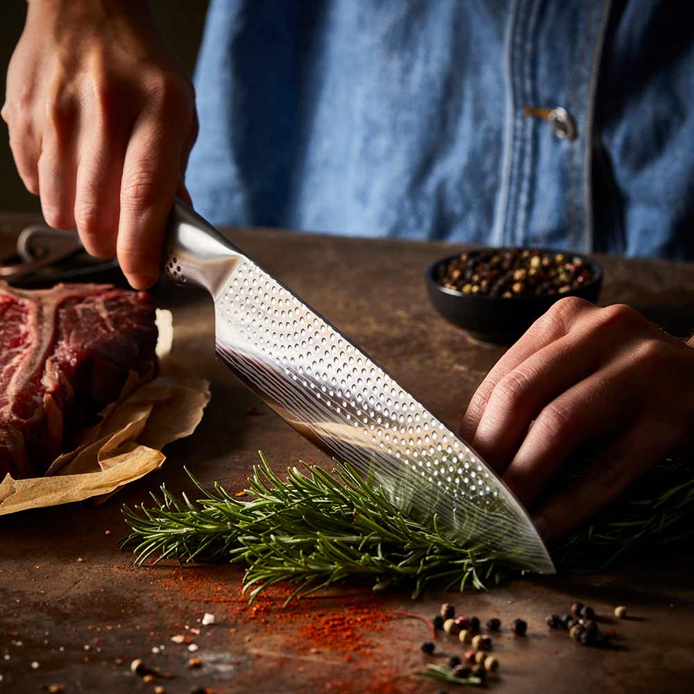 A person chops herbs in a kitchen with large steak in background on a wooden board - Baccarat