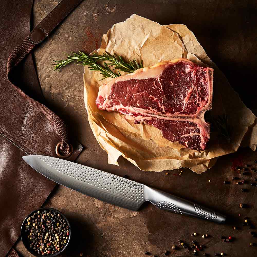 A person chops herbs in a kitchen with large steak in background on a wooden board - Baccarat