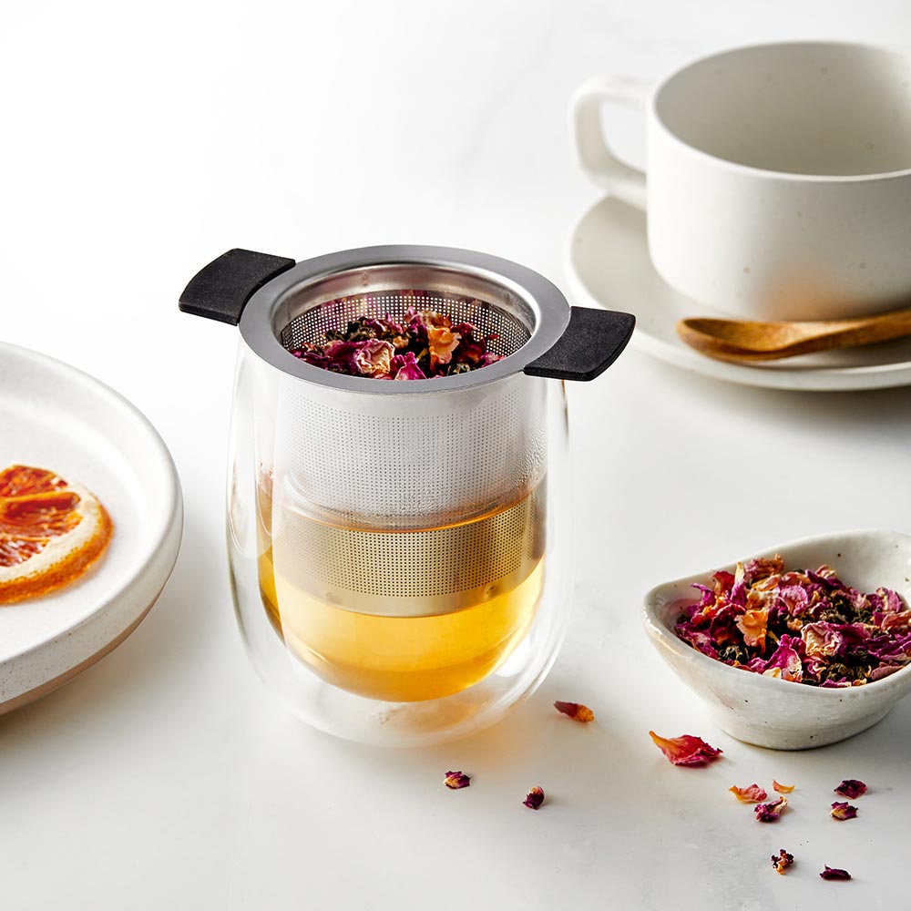glass teacup with metal infuser golden tea and rose petals next to a white teacup - Baccarat
