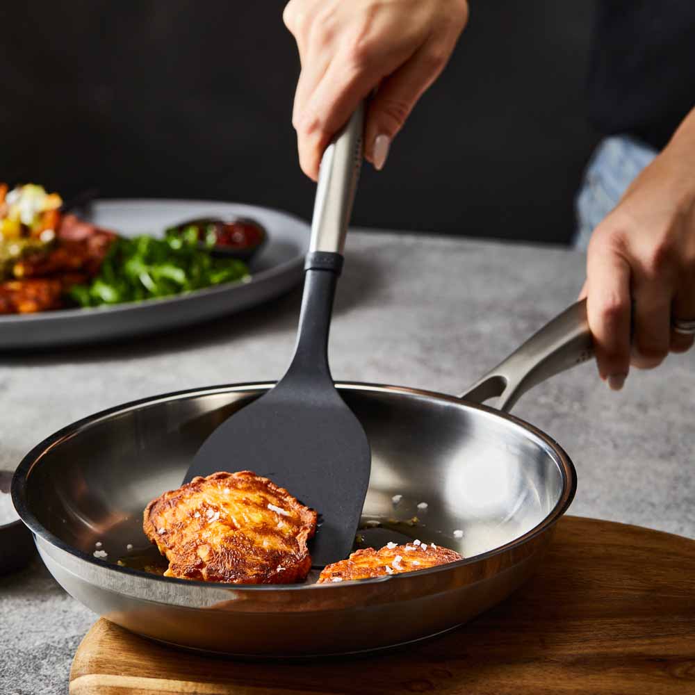 Person flips chicken with a black spatula in a stainless steel frying pan on wooden board - Baccarat