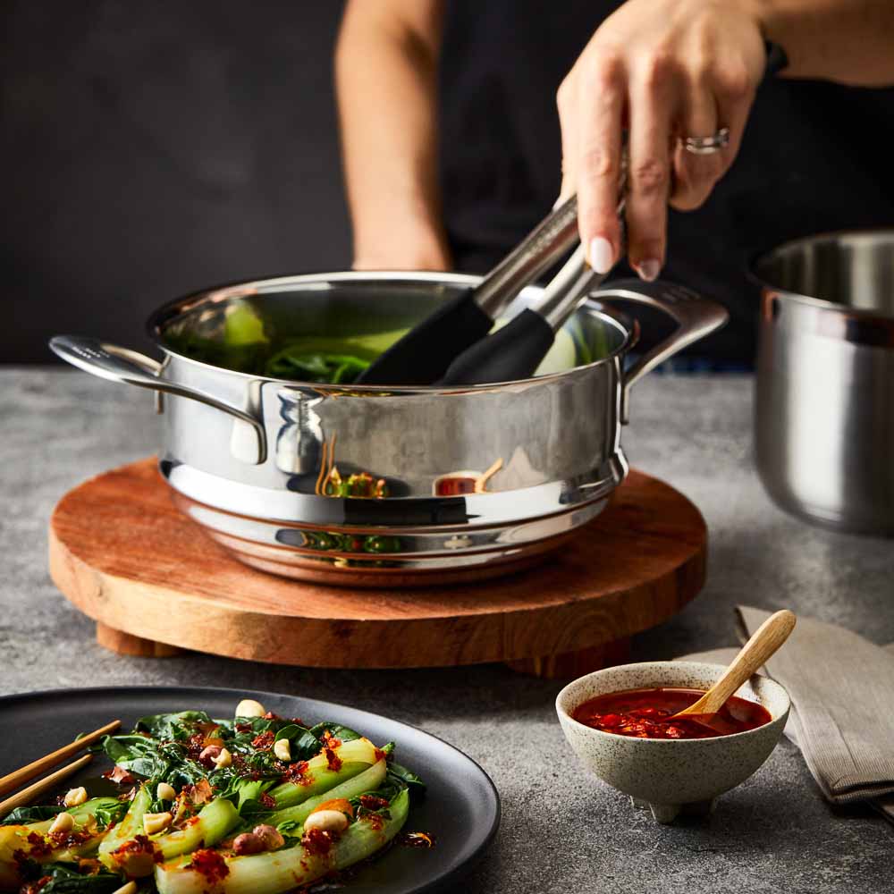 Hand lifting lid off stainless steel steamer with green vegetables on wooden board 32cm - Baccarat
