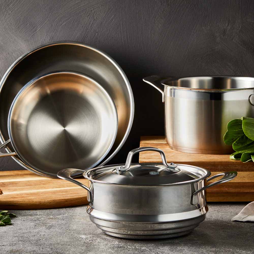 Four stainless steel cookware pieces on a gray surface including pots and pans with lid - Baccarat