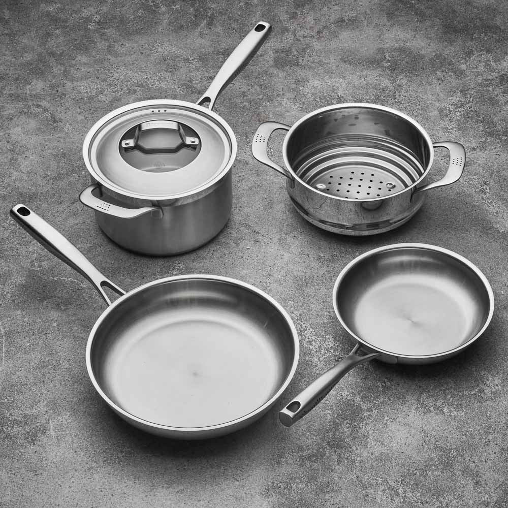 Four stainless steel cookware pieces on a gray surface including pots and pans with lid - Baccarat