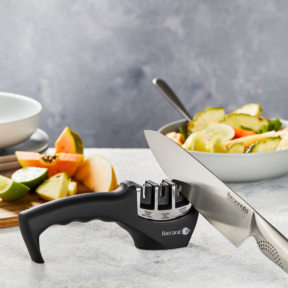 A person sharpens a large knife on a kitchen counter beside fruits- Baccarat 3-Step Knife Sharpener