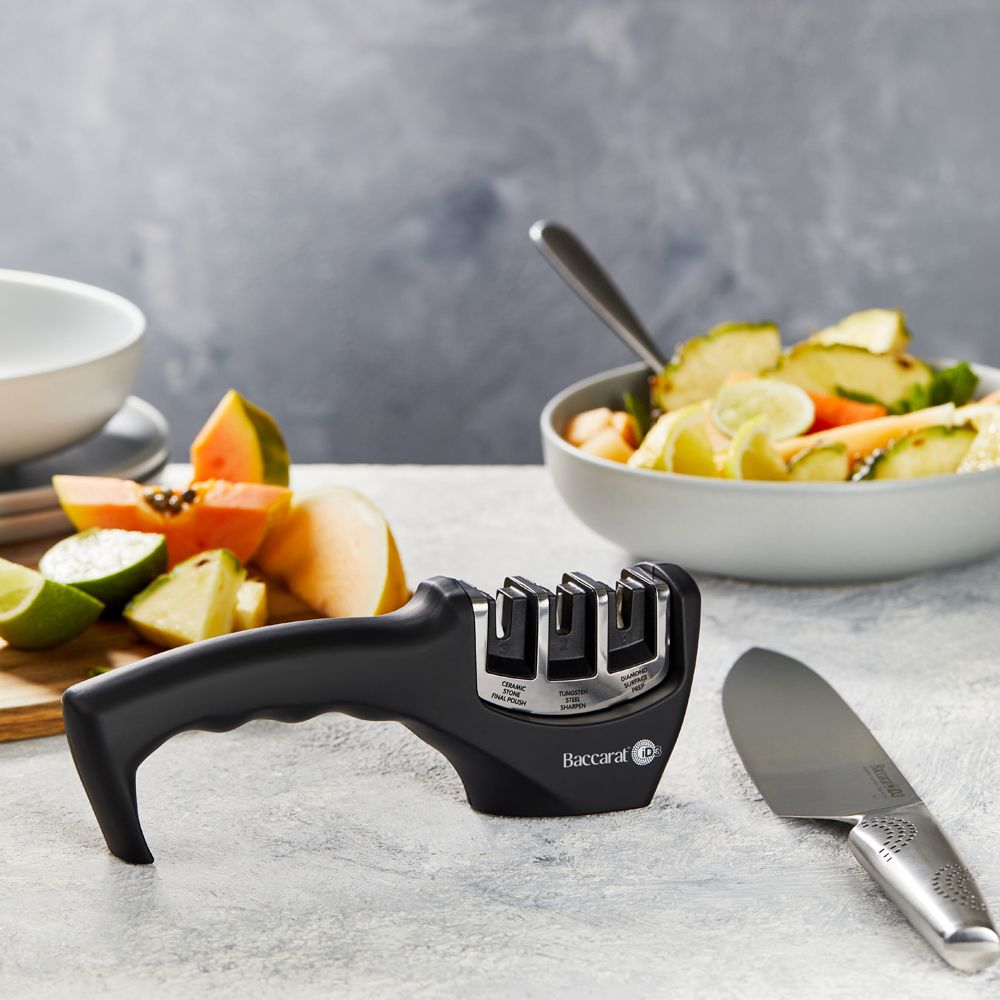 A person sharpens a large knife on a kitchen counter beside fruits- Baccarat 3-Step Knife Sharpener