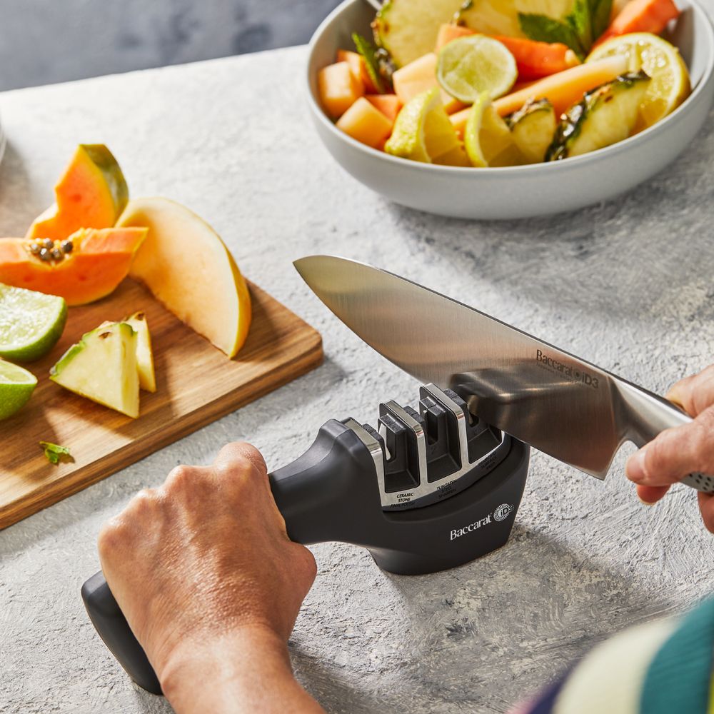 A person sharpens a large knife on a kitchen counter beside fruits- Baccarat 3-Step Knife Sharpener