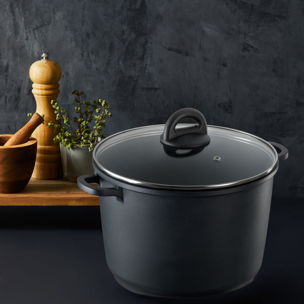 A matte dark gray stockpot with a matching dark gray glass lid and black handle, shown on a dark studio background with a wooden pepper mill, mortar, and small plant as props.