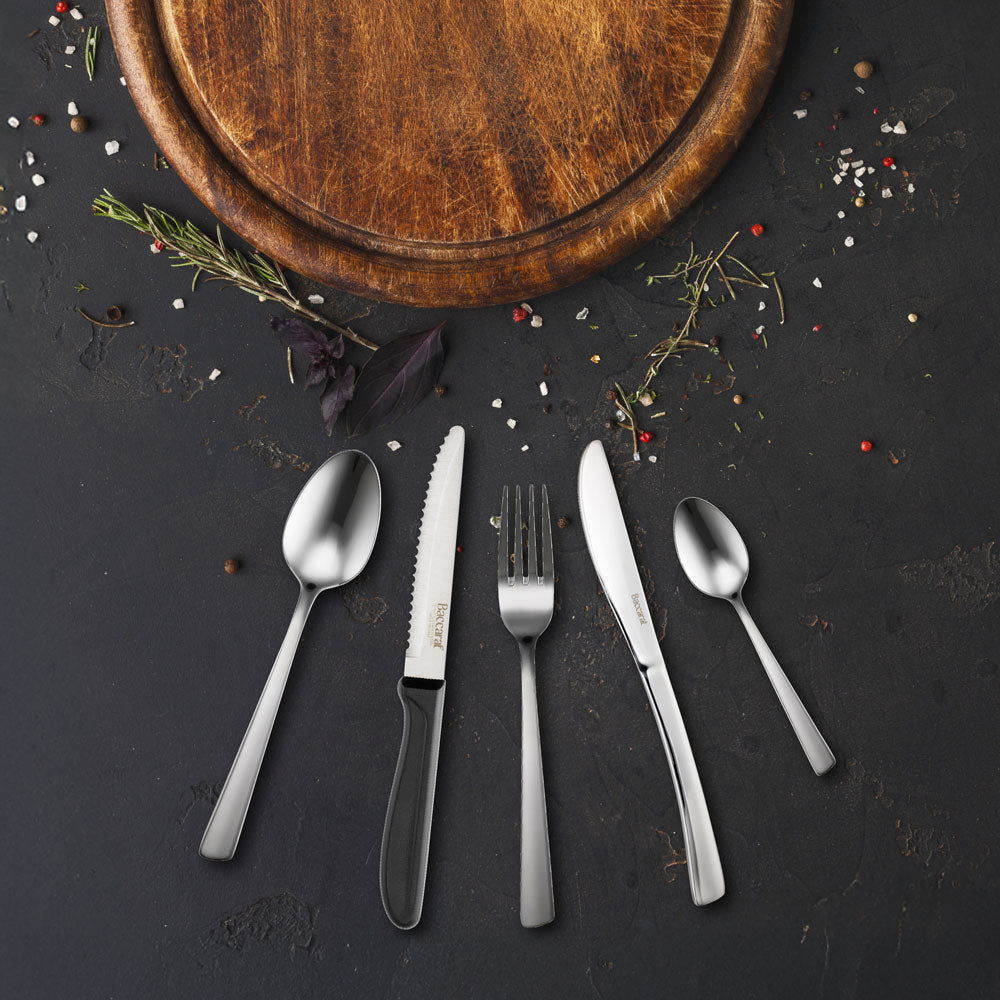 A five-piece stainless-steel cutlery set arranged on a dark, textured surface, with a wooden cutting board, herbs, and spices as styling props. The set includes a dinner fork, a salad fork, a dinner spoon, a dinner knife, and a salad spoon, with the dinner fork and knife featuring black handles.