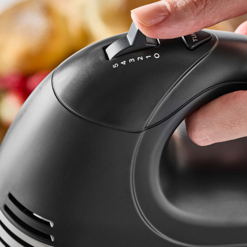 A hand is adjusting the speed control switch on top of the black hand mixer.