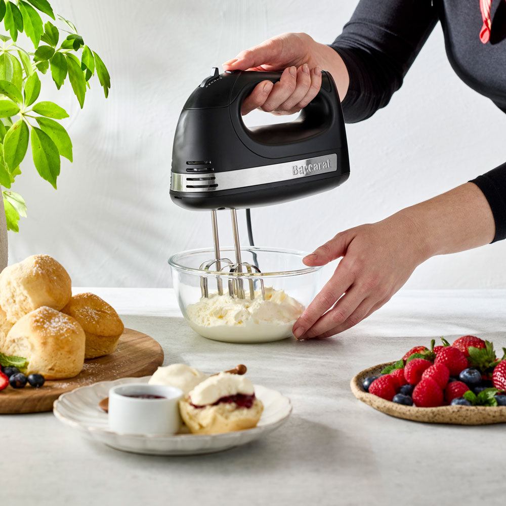 A person's hands are using the black hand mixer to whip cream in a glass bowl on a kitchen table.