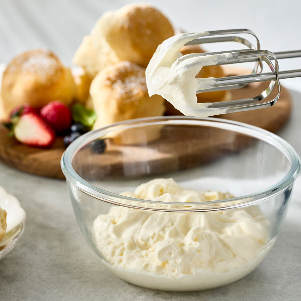 Whipped cream is on the beaters of a hand mixer hovering over a glass bowl next to freshly baked scones and berries.