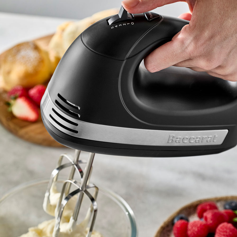 A close-up view shows a hand holding and operating the black hand mixer while mixing ingredients in a bowl.