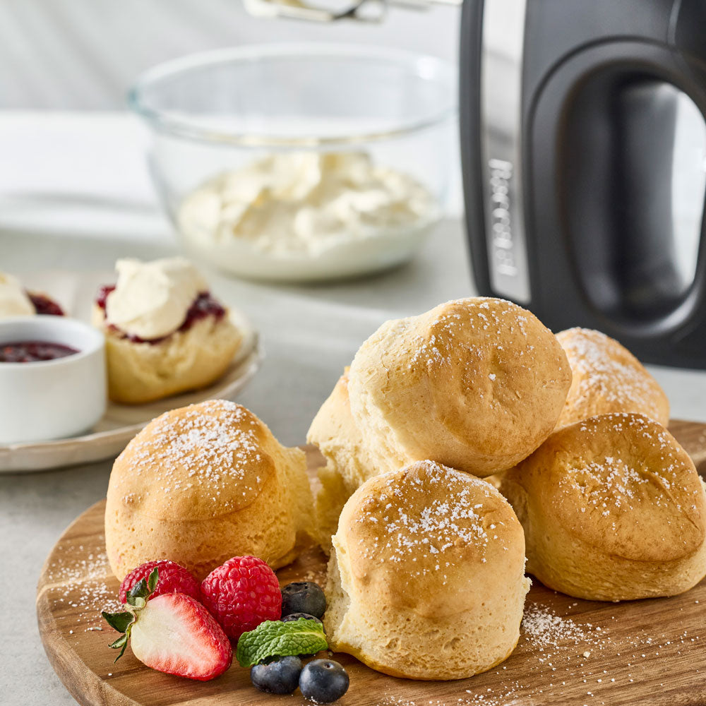Several freshly baked scones are dusted with powdered sugar and arranged on a wooden board next to a bowl of cream and berries.