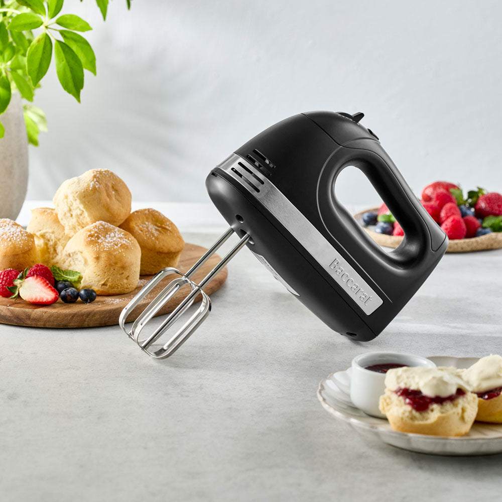 A black hand mixer is propped up on a counter next to scones, jam, and berries on a plate.