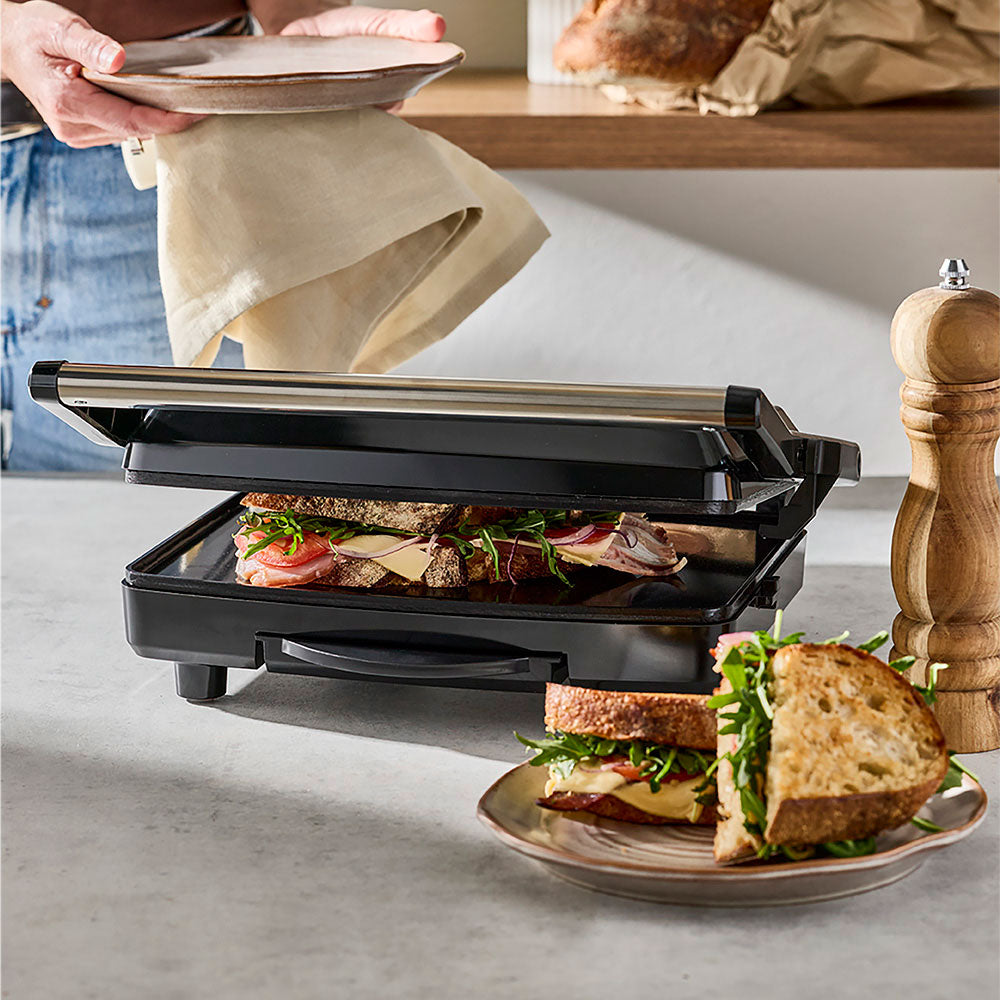 A lifestyle shot shows the sandwich maker in use on a kitchen counter with finished sandwiches on a plate.