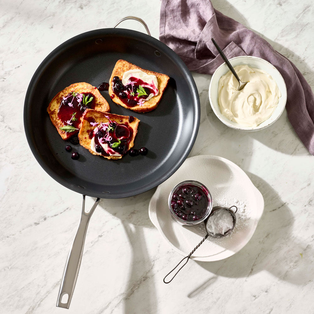 French toast topped with berries and cream is arranged in a black pan alongside small bowls on a table.