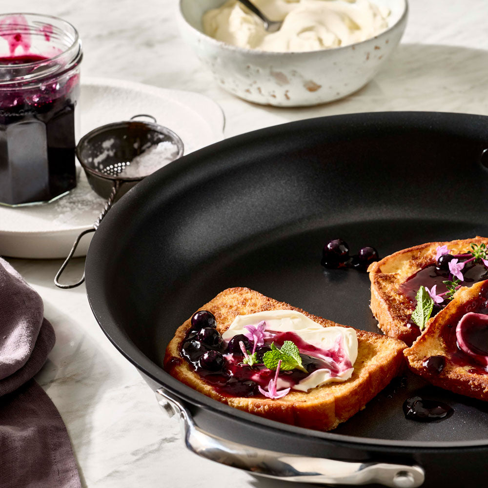 A close-up view focuses on French toast with berry topping inside a dark pan next to a jar of jam.