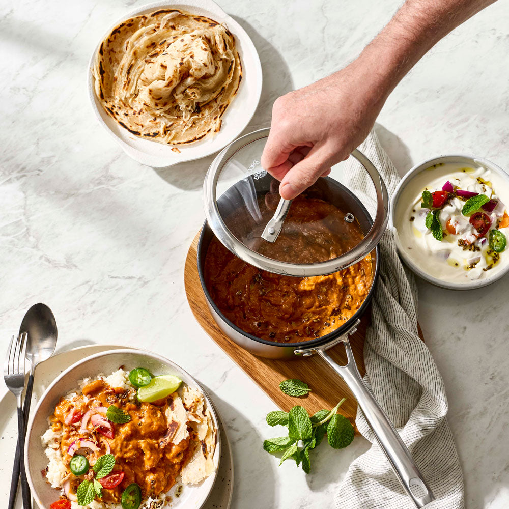 A hand lifts a glass lid from a stainless steel saucepan containing an orange-brown curry, set on a wooden board with a folded cloth. Surrounding plates include flatbread, a white sauce with fruit and mint, and another plated curry with lime and onions, all arranged on a light marble surface.