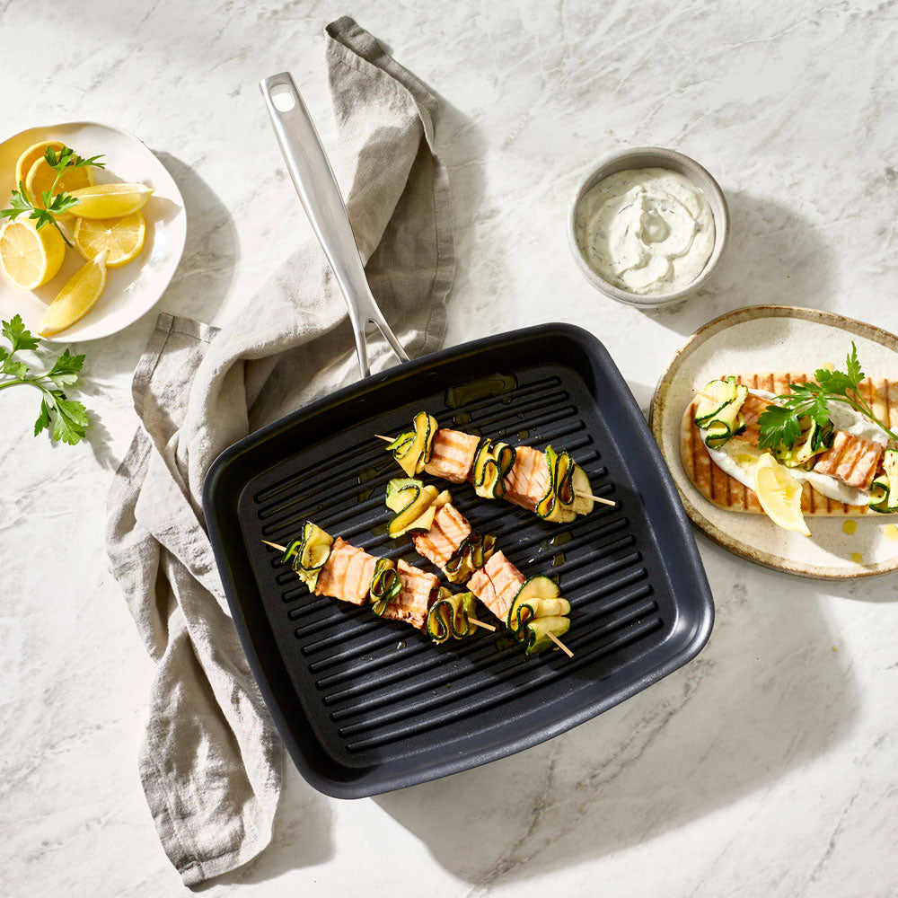 An overhead shot shows food cooking in a square grill pan surrounded by dishes and a cloth napkin.