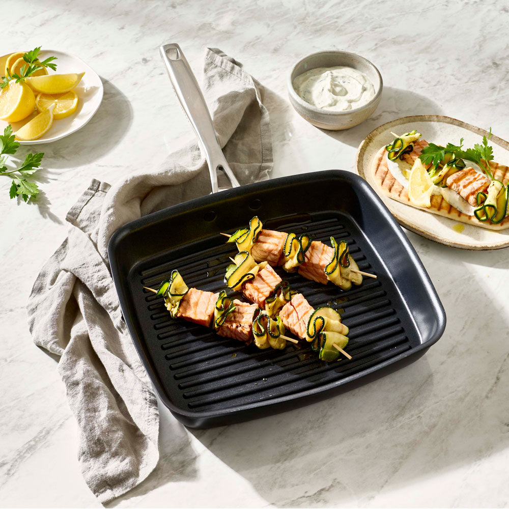 Skewered food is cooking inside a square grill pan on a tabletop setting.