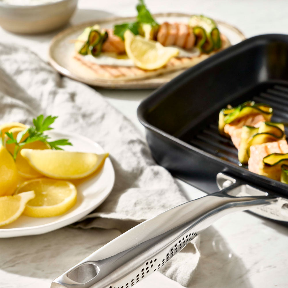 A grill pan is positioned on a table next to a plate with lemon wedges and wrapped food.
